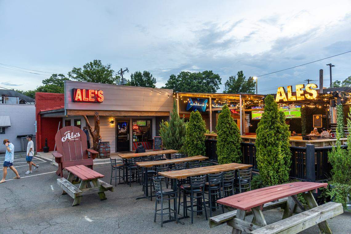 Exterior view of a bar and restaurant called “Ale’s” at dusk, featuring an outdoor patio area. The building on the left has a gray facade with a prominent red sign that says “ALE’S.” In front of it is a large, wooden Adirondack chair decoration painted red, next to a picnic table. To the right is an elevated outdoor seating area with string lights and a large sign spelling out “ALES” in illuminated yellow letters. The patio has several tall wooden tables with black stools and a few red picnic tables in the foreground. People are visible walking on the left and sitting on the elevated patio. The sky is overcast.