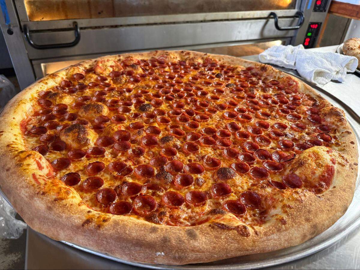 An eye-level, close-up shot of a massive, freshly baked New York-style pepperoni pizza resting on a large circular metal tray. The pizza is densely covered from edge to edge with small, crispy cup-and-char pepperoni slices over bubbly melted mozzarella cheese and red sauce. The thick crust is golden-brown with slight charring and large air bubbles. In the blurred background, a stainless-steel commercial pizza oven is visible.