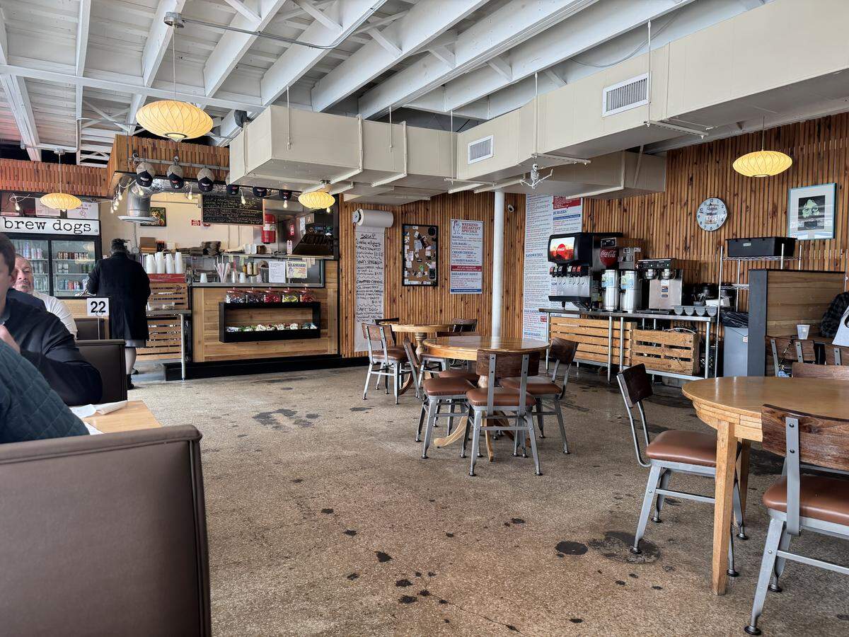 The interior of a spacious restaurant with exposed white ceiling beams and wood-paneled walls. Circular wooden tables and booths are spread across a concrete floor, with a drink station and service counter in the background.