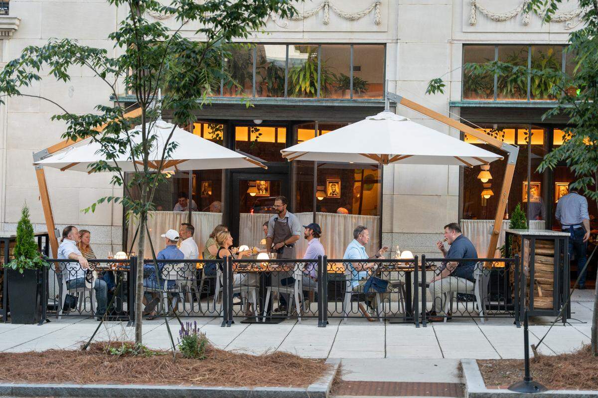 Patrons dine at an upscale outdoor patio area in front of a stone building. Several small tables are occupied by groups of people, shaded by two large, rectangular white patio umbrellas with light wood frames. A waiter in a gray apron stands near a central table, and glowing orb-like lamps sit on the tables as evening sets in. Large windows reveal a warmly lit interior with greenery and framed artwork, while the exterior is defined by a low black decorative fence and young trees in the foreground.