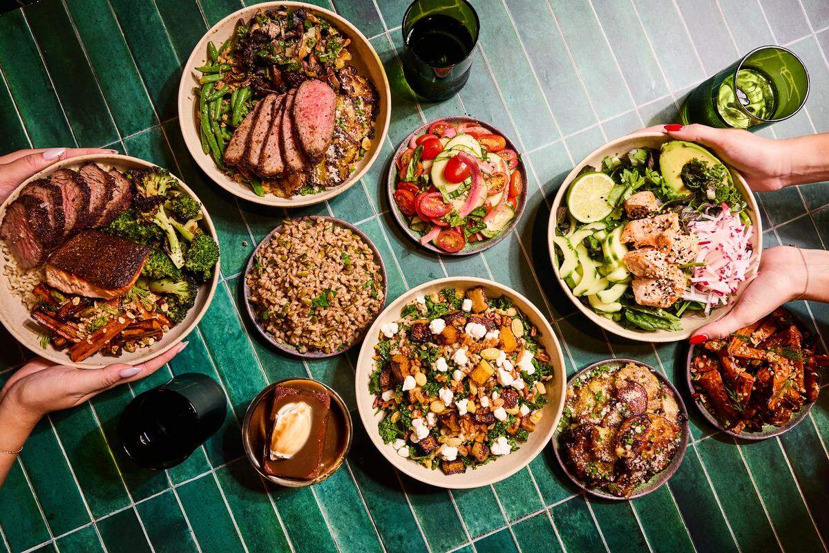 A top-down overhead shot of a vibrant spread of various healthy bowls and side dishes arranged on a green-tiled table. The meal includes bowls featuring sliced medium-rare steak, seared salmon, roasted broccoli, and grains. Centered are smaller plates of tomato and cucumber salad, roasted sweet potatoes, and a kale salad with feta and squash. Two pairs of hands are visible on the left and right, reaching for the bowls, and a small dessert with caramel sauce and cream sits at the bottom.