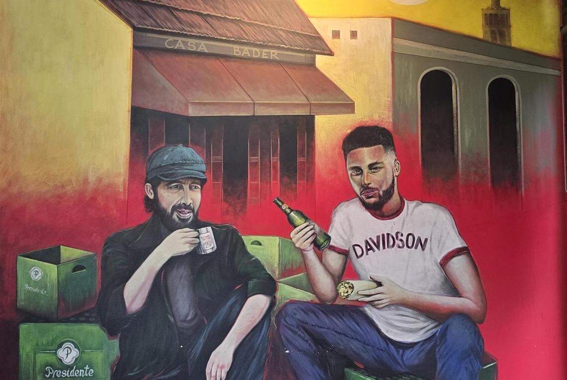 A detailed painting on a wall shows two men sitting on green Presidente beer crates in front of a yellow building labeled ‘CASA BADER’. The person on the right, wearing a “Davidson” T-shirt, holds a bottle and a small sandwich. The person on the left, in a dark cap, holds a white mug.