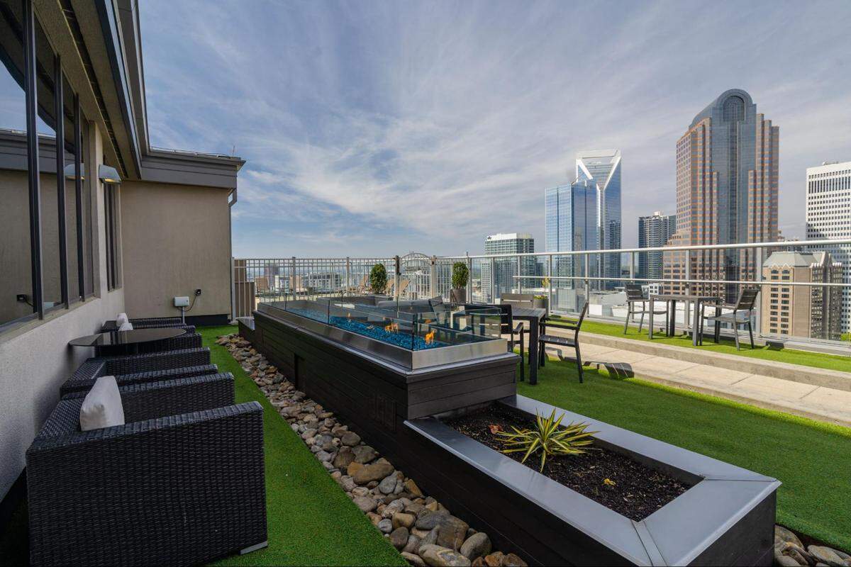 A panoramic shot of a modern rooftop patio with a view of a city skyline. The patio features a long, rectangular, dark wood structure with an embedded planter box and a glass-enclosed water feature or fire pit. To the left, a row of dark wicker lounge chairs with white cushions is visible. The ground is a mix of artificial green grass, river stones, and paving. In the background, multiple tables and chairs are set up, all against a backdrop of tall city buildings and a cloudy blue sky. A tall skyscraper with a unique jagged top is prominent in the skyline.