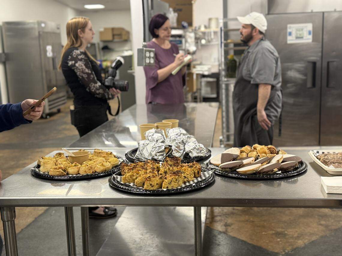 A wide shot of a professional kitchen during a media event or interview. In the foreground, a stainless steel table holds platters of kugel, black-and-white cookies, and wrapped sandwiches. In the background, a chef talks to a reporter taking notes while another person holds a professional camera with a flash.