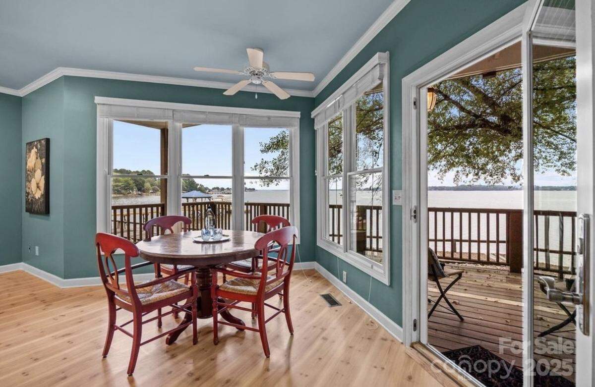 A bright dining or breakfast nook with teal walls and a light blue ceiling, featuring a circular wooden table surrounded by four red wooden chairs. The room has light wood floors and is enclosed by large windows and a glass sliding door that offer a stunning, expansive view of a lake and a surrounding wooden deck and railing.
