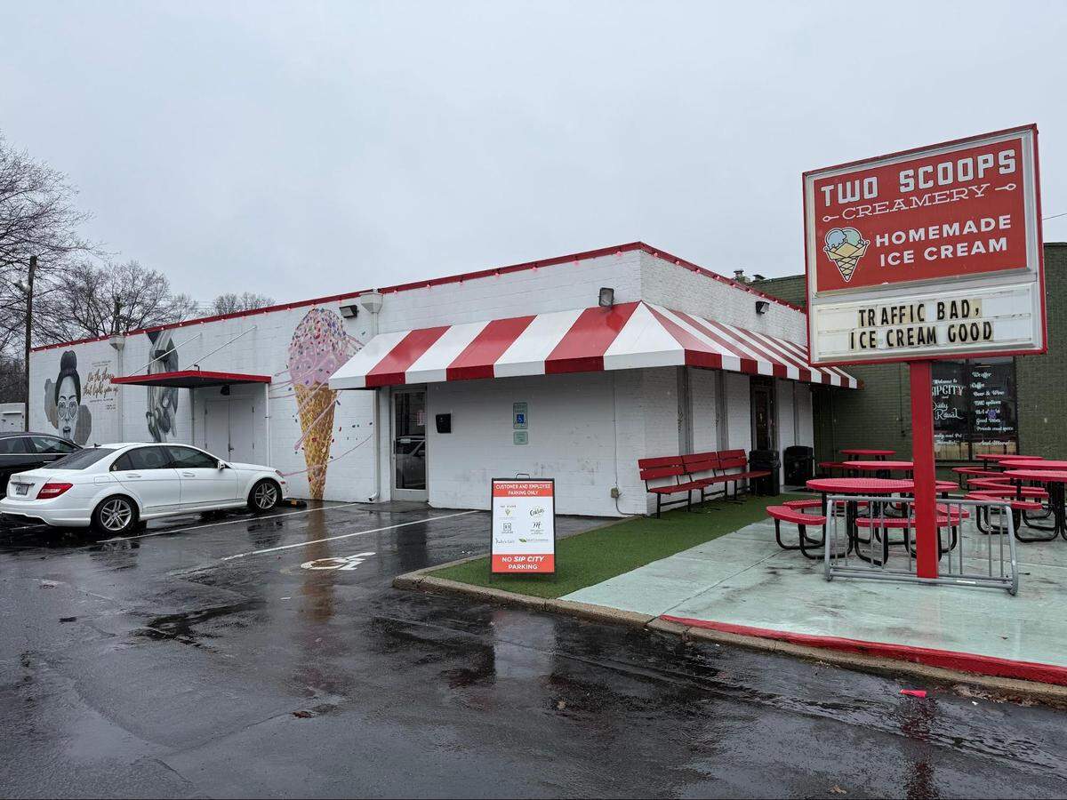 The exterior of a white brick building with a red-and-white striped awning. A large red sign features the “Two Scoops Creamery” logo and a marquee that reads, “TRAFFIC BAD, ICE CREAM GOOD.” The parking lot is wet from rain, and several red outdoor picnic tables are visible.