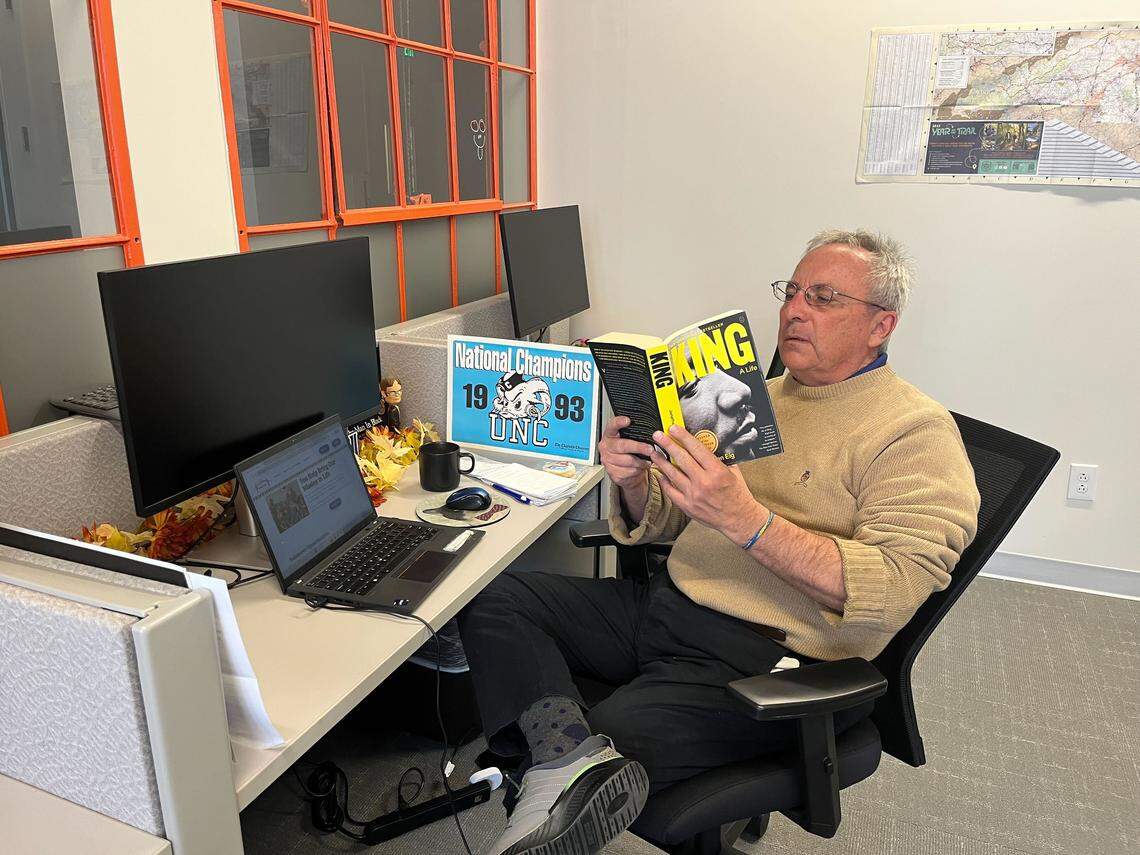 A person in a tan sweater and glasses sits at a desk in an office cubicle, reading a biography of Martin Luther King Jr. titled “King.” A laptop, two monitors, and a “National Champions 1993 UNC” sign are also on the desk.