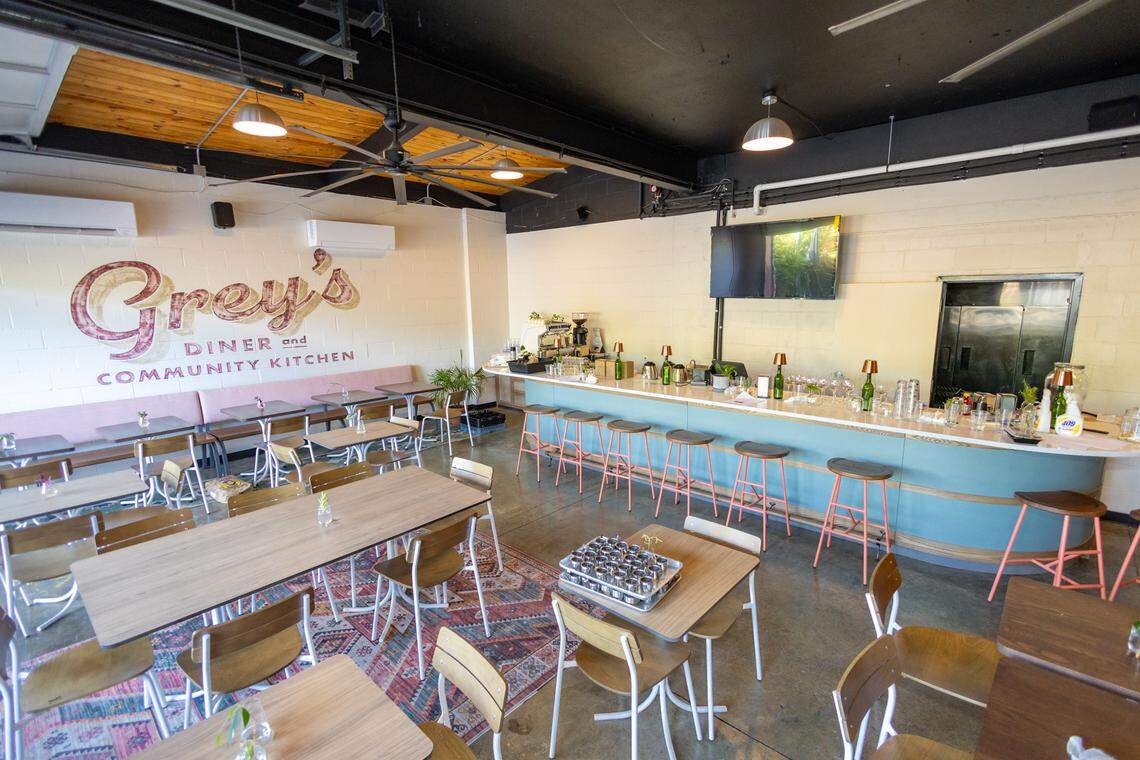 A wide interior view of the modern “Grey’s Diner and Community Kitchen” dining room. The space features a polished concrete floor, a large hand-painted mural on a white cinder block wall, and a curved light blue service counter with wooden-topped stools. Various wooden tables and chairs are spread throughout the room, some resting on a colorful geometric area rug. A high industrial ceiling with a large wooden-bladed fan and pendant lights completes the bright, open atmosphere.