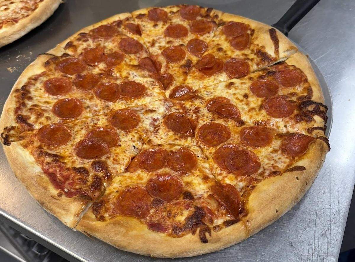 A high-angle shot of a freshly baked, round pepperoni pizza on a metal pizza peel. The pizza is sliced into eight pieces and covered with a generous amount of melted cheese and slightly curled, crispy pepperoni slices. The crust is thick and golden-brown. Another pizza is partially visible in the upper left corner, also on a metal surface.