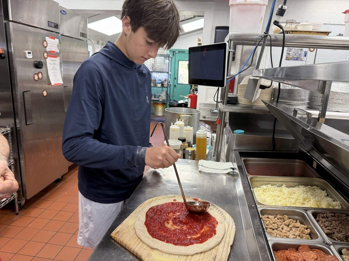 A person wearing a dark blue hoodie is preparing a pizza in a commercial kitchen. They are using a metal ladle to spread red tomato sauce in a circular motion over a stretched pizza dough resting on a wooden peel. To the right, there is a stainless steel prep station with built-in refrigerated containers holding various pizza toppings, including shredded cheese, crumbled sausage and pepperoni.