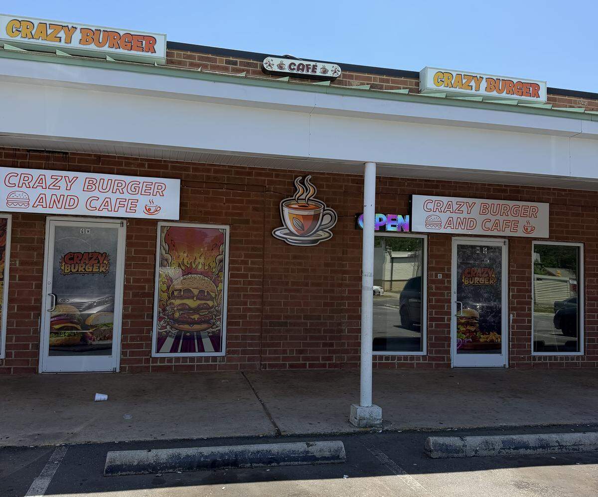 An eye-level outdoor shot of the storefront for “Crazy Burger and Cafe,” located in a strip mall. The building features a red brick facade with a wide, white overhang supported by a central white support column. Multiple signs advertise the business: two rectangular signs on the roofline read “CRAZY BURGER” in orange letters with a green border, flanking a small oval “CAFÉ” sign. Below the overhang, two identical white banners feature orange text that reads “CRAZY BURGER AND CAFE” with small line-art icons of a burger and a coffee cup. Between the two main glass entrance doors, a large, stylized graphic of a steaming coffee cup is mounted directly onto the brick wall.
