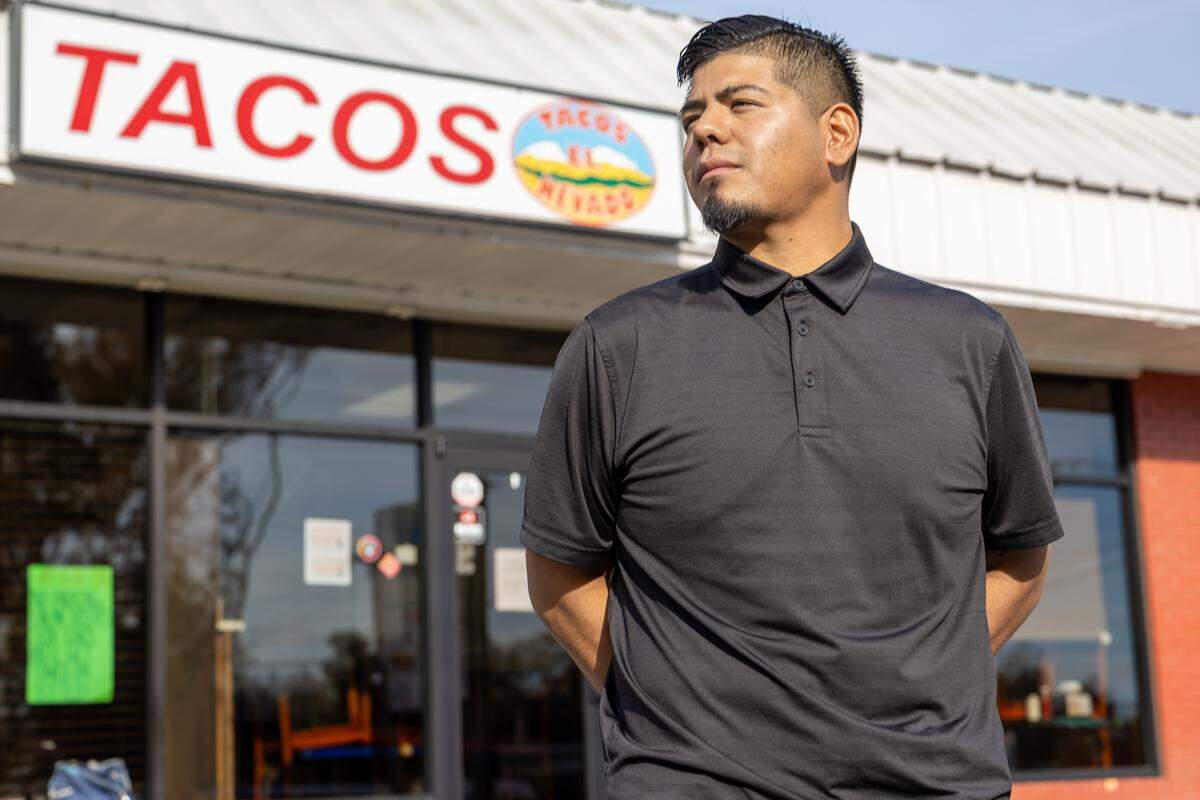 A medium, eye-level portrait of a restaurant worker wearing a black polo shirt, standing with hands clasped behind their back and a serious expression. The worker is standing in front of a glass storefront, with a sign overhead prominently reading “TACOS” in red letters.