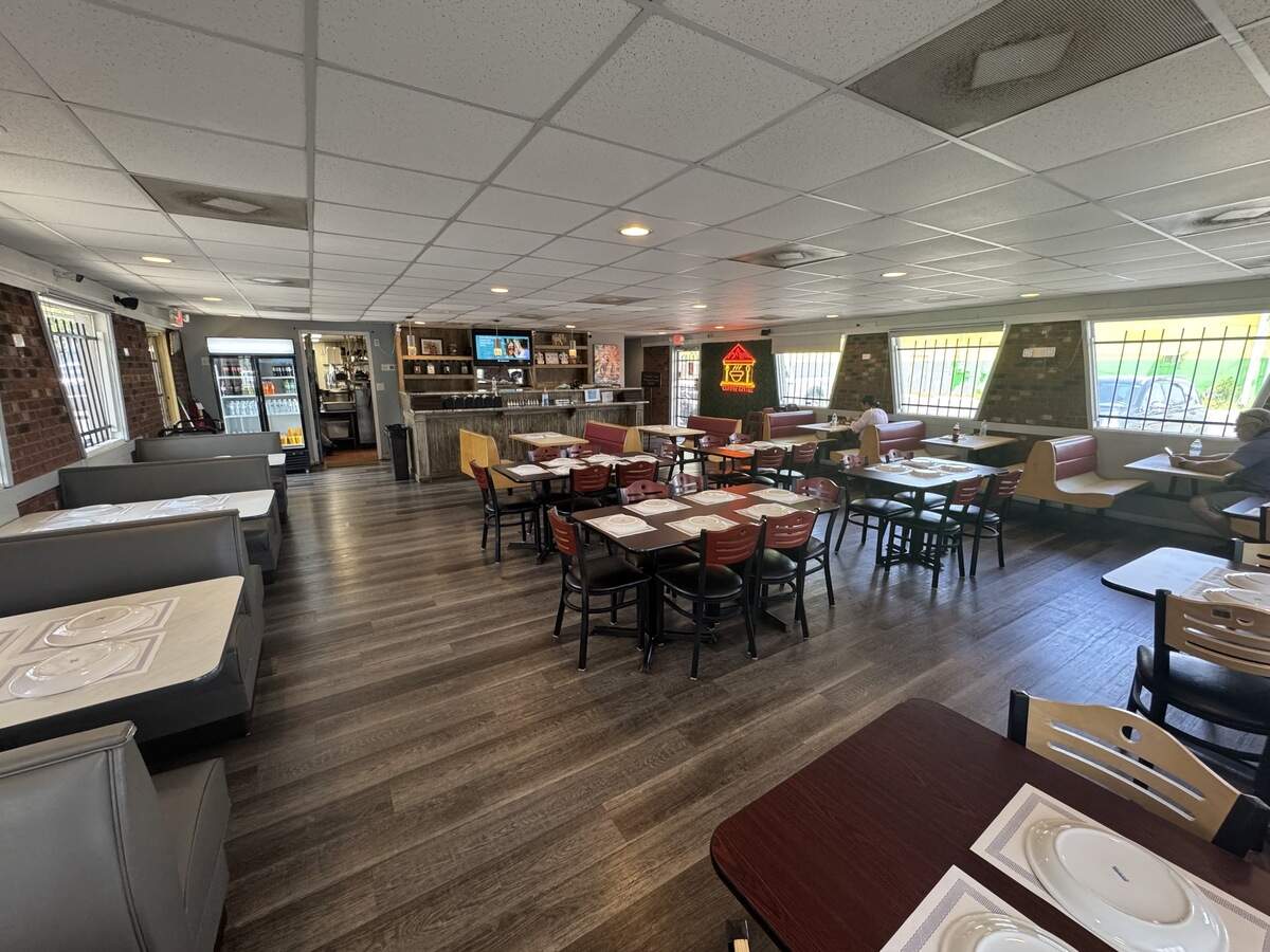 A wide-angle view of the spacious interior of the restaurant. The dining room features wood-look flooring, multiple rows of gray booths along the left wall, and dark wood tables with reddish-brown chairs arranged in the center. Large, slanted windows line the right wall, letting in daylight. In the back, a wooden service counter sits below a TV, and a glowing neon sign shaped like a house is visible on a far wall.