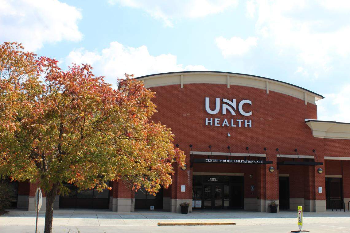 The exterior of a brick UNC Health building on a sunny day. The large white letters ‘UNC HEALTH’ are visible above the entrance, with smaller text reading ‘CENTER FOR REHABILITATION CARE’ and the address ‘1907’. An autumn tree with red and green leaves partially obscures the left side of the building.