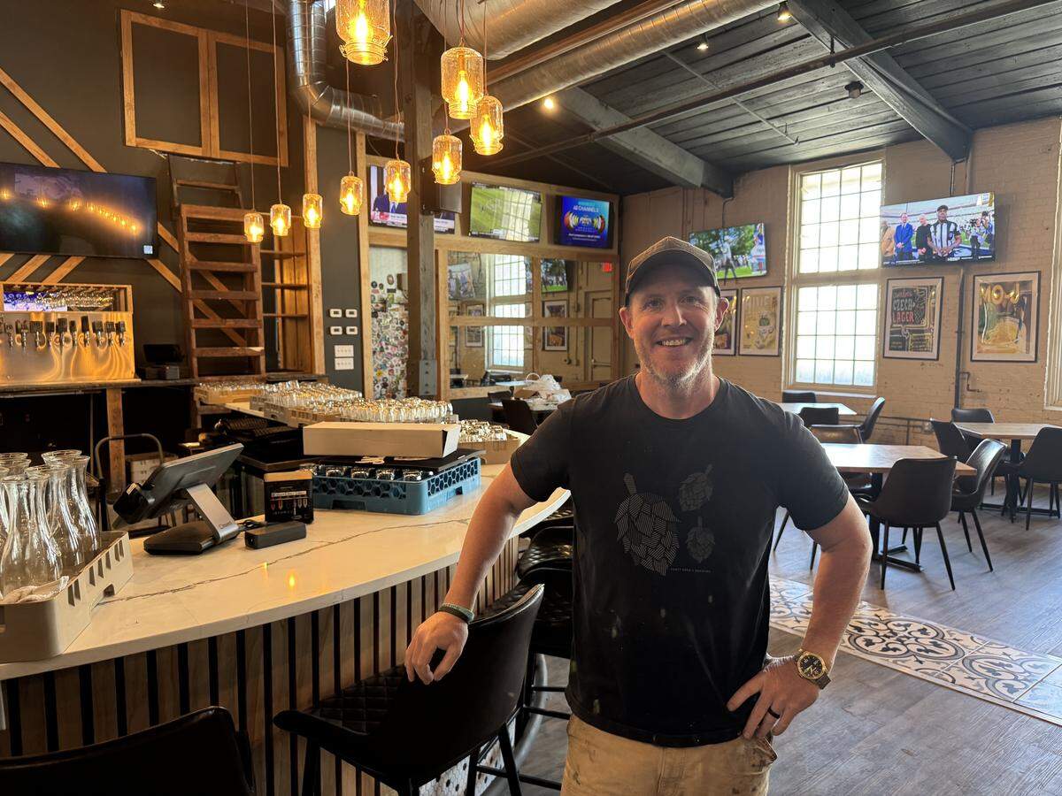A medium shot of a smiling man wearing a black graphic T-shirt and a baseball cap, standing inside the brewery. He is leaning against the curved white bar top. The background shows the warmly lit interior, including the Edison bulb lighting, a tap wall, and the dining area with wooden tables and dark chairs. The atmosphere is bright and inviting.