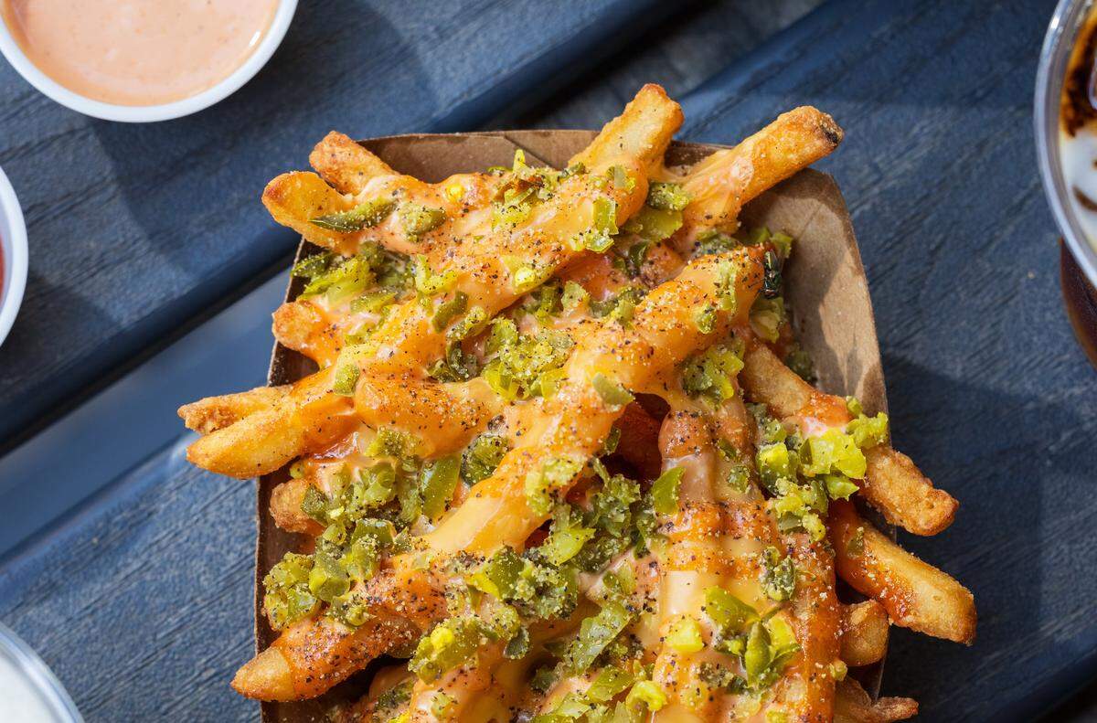 On a dark blue surface under bright sunlight, a paper boat is overflowing with “Beast Mode Fries.” The thick-cut fries are smothered in a cheese sauce, covered with a layer of green jalapeño or pickle relish, and seasoned with black pepper.