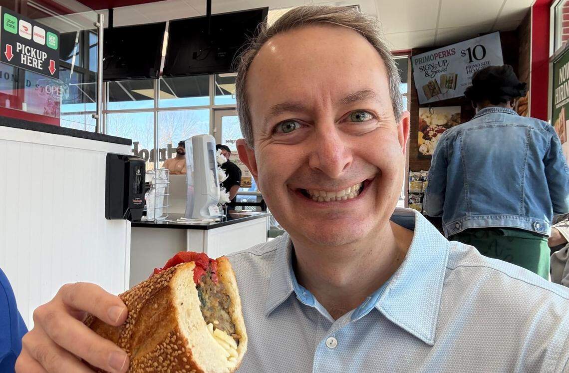 A selfie of a smiling person in a light blue polo shirt holding a large hoagie sandwich with a sesame seed roll. The background shows the bright interior of a PrimoHoagies, including a “Pickup Here” sign and a person in a denim jacket.