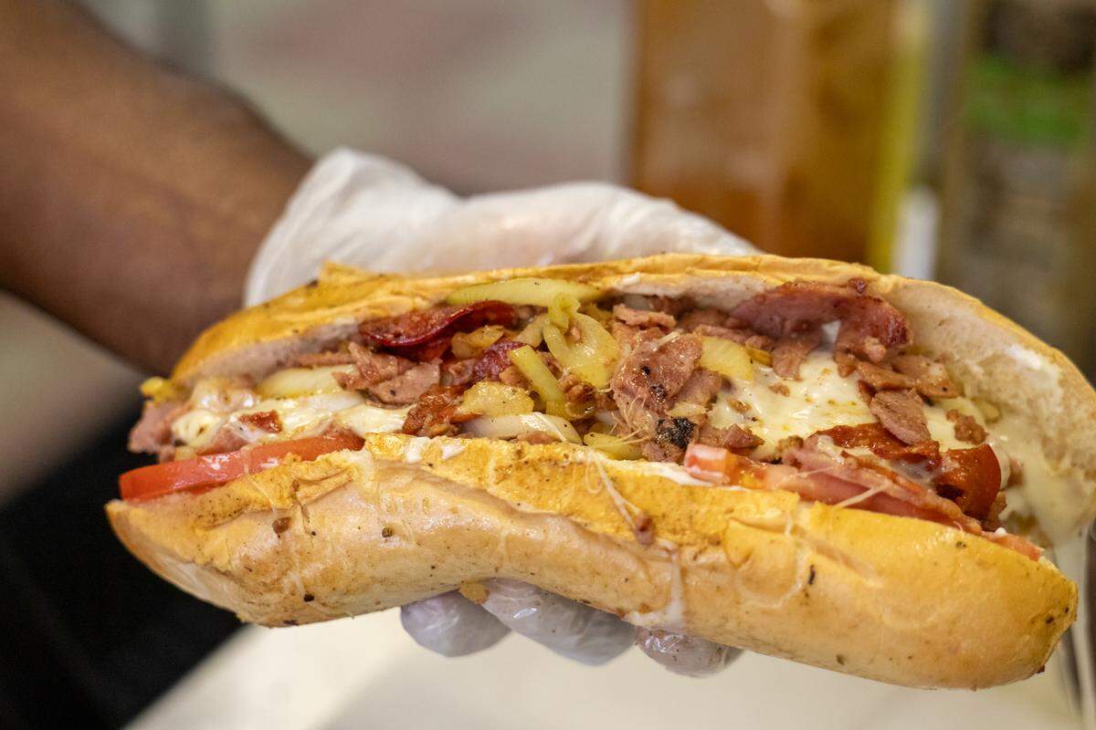 A close-up, eye-level shot of a gloved hand holding a large, loaded sub sandwich. The sandwich features melted white cheese, grilled meats, pepperoni, sliced tomatoes, and onions, all spilling out of a toasted hoagie roll.