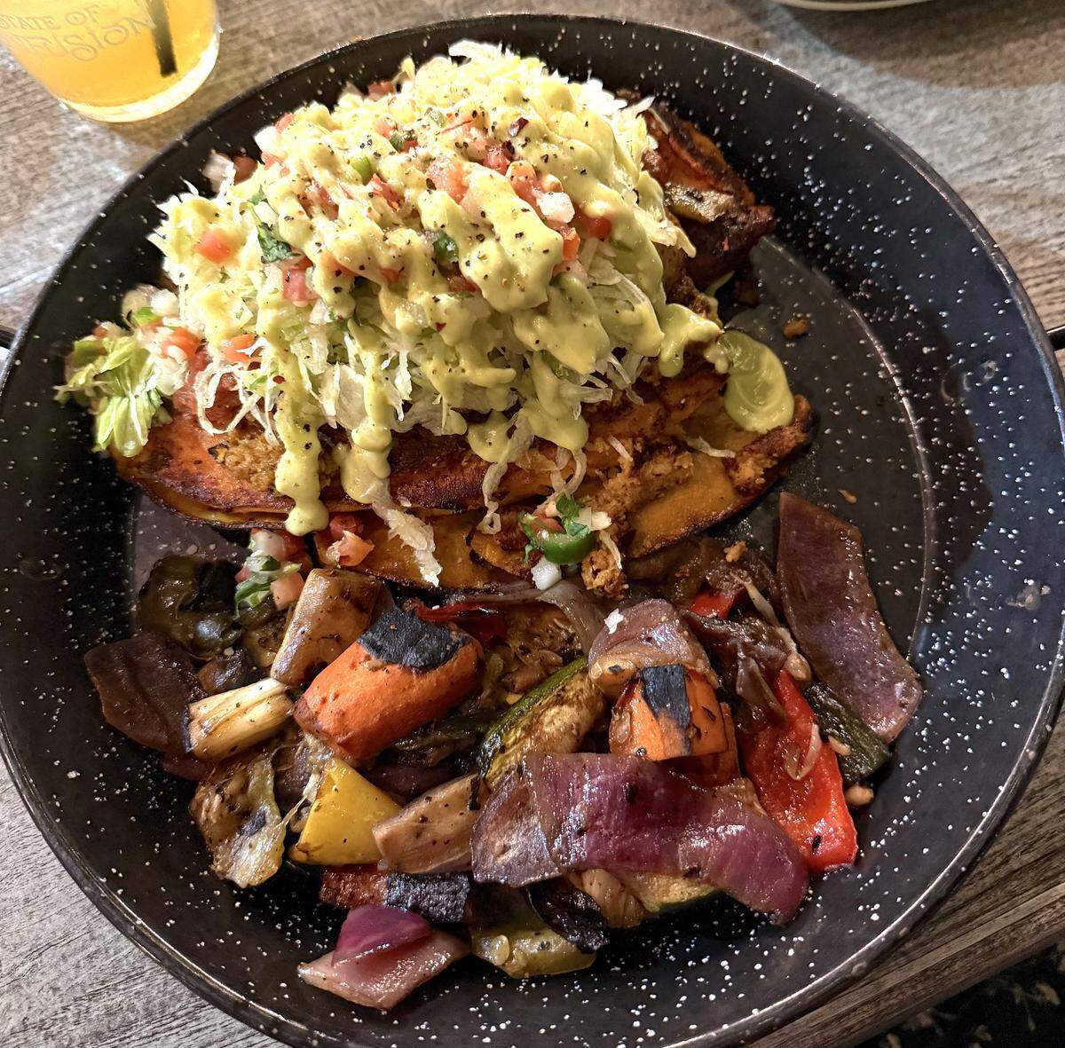 A top-down view of a rustic meal served in a black, white-speckled enamel skillet on a wooden table. The top half of the skillet contains a large, crispy, folded tortilla or flatbread that is piled high with shredded lettuce, pico de gallo, and generously drizzled with a creamy, light green avocado sauce. The bottom half of the skillet is filled with a hearty portion of chunky, roasted vegetables featuring visible char marks, including red onions, carrots, zucchini, and bell peppers. In the top left corner, the base of a glass filled with a yellow beverage and a straw is partially visible, bearing the faint text “STATE OF CONFUSION”.
