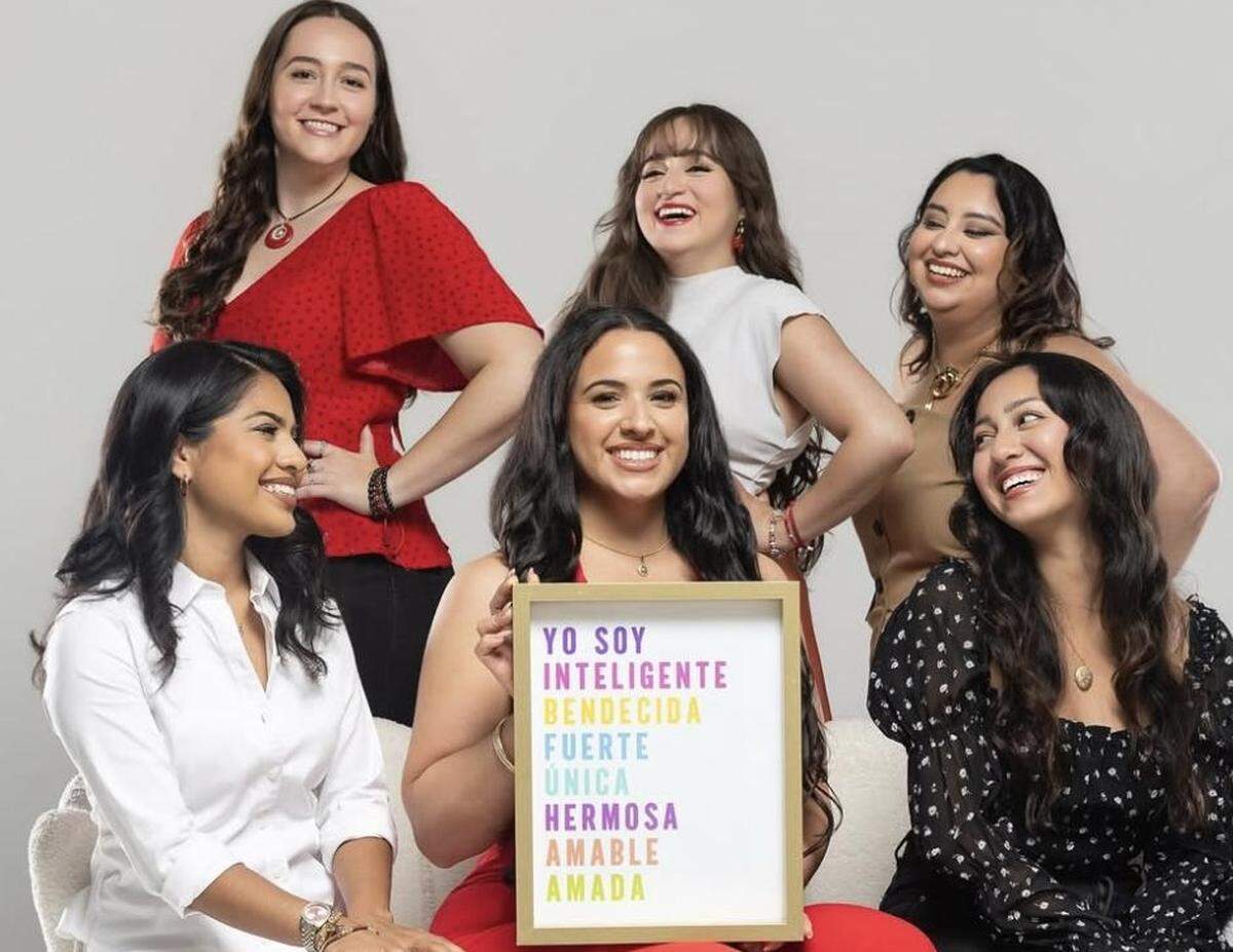 A group studio portrait of six smiling women of diverse backgrounds. The central figure holds a framed sign with a motivational message in Spanish written in rainbow text: “YO SOY INTELIGENTE BENDECIDA FUERTE ÚNICA HERMOSA AMABLE AMADA.”