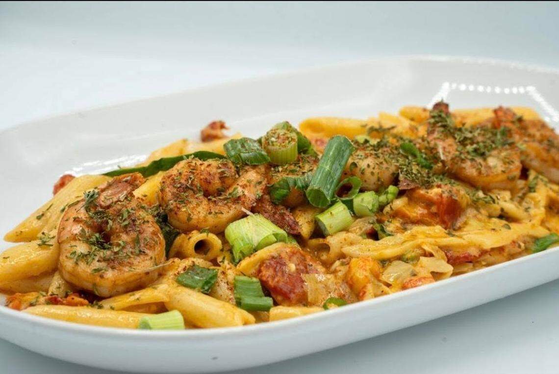 A close-up of penne pasta tossed in a creamy, orange-tinted sauce, topped with several blackened grilled shrimp. The dish is heavily garnished with sliced green onions and dried herbs, served on a long, white oval platter