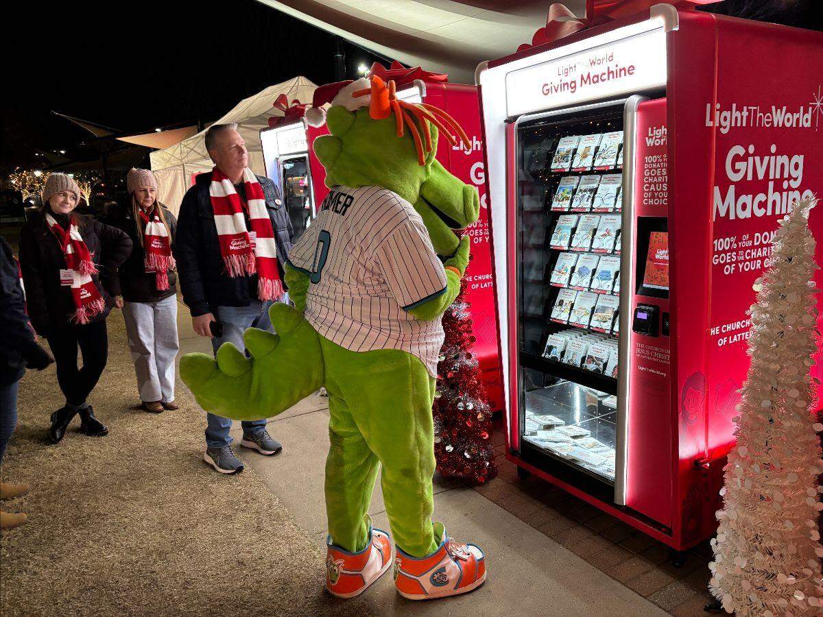 A person in a green dragon mascot costume and a few people in scarves stand near a large red “LightTheWorld Giving Machine,” which resembles a vending machine.