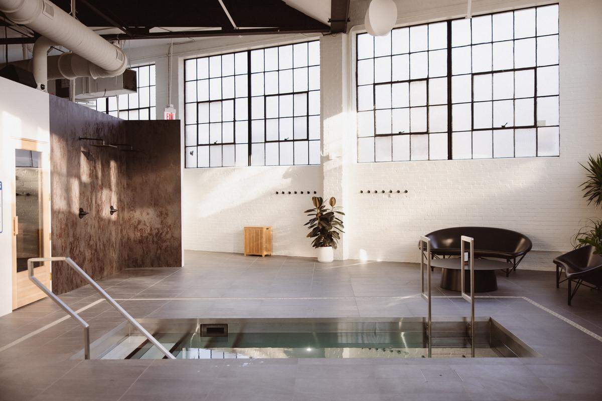 A wide shot of a large, modern wellness studio with high ceilings and industrial-style windows. In the foreground, a metal-lined cold plunge pool is built into the floor, featuring metal handrails for entry. To the left, a textured, dark grey wall holds two open-concept showerheads.
