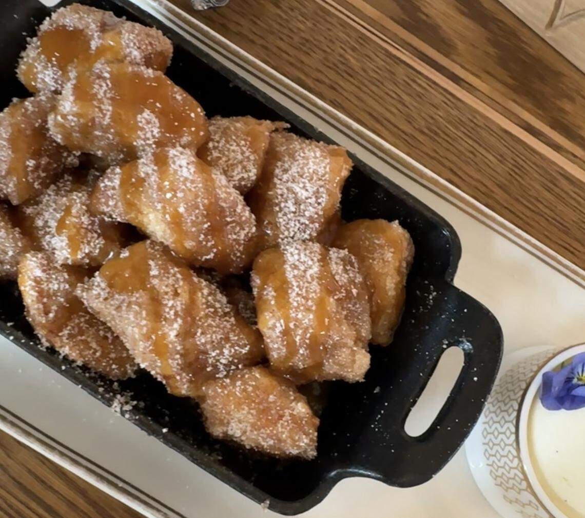 A tempting close-up of pull-apart monkey bread served in a rustic, black cast-iron dish. The bite-sized pieces of dough are golden-brown, heavily coated in granulated cinnamon sugar, and generously drizzled with a glistening caramel sauce.