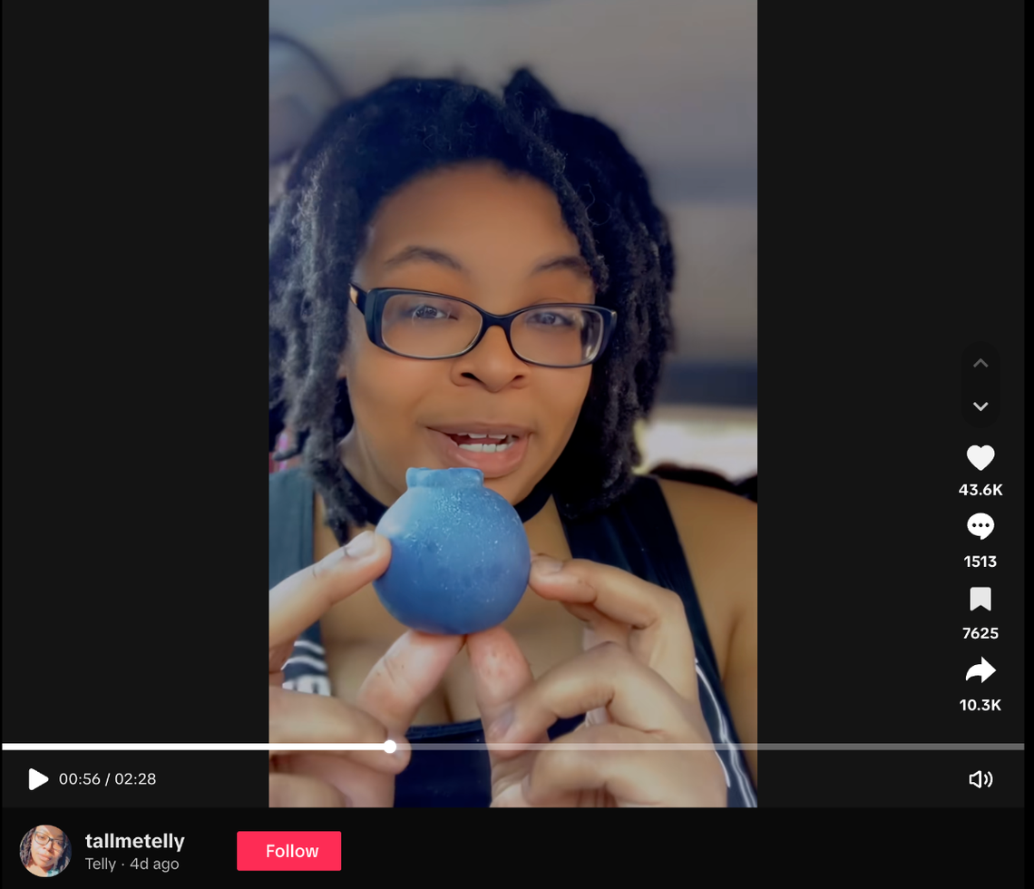A screenshot of a TikTok video by user “tallmetelly.” A woman with glasses and locs holds up a small, realistic blueberry-shaped pastry.