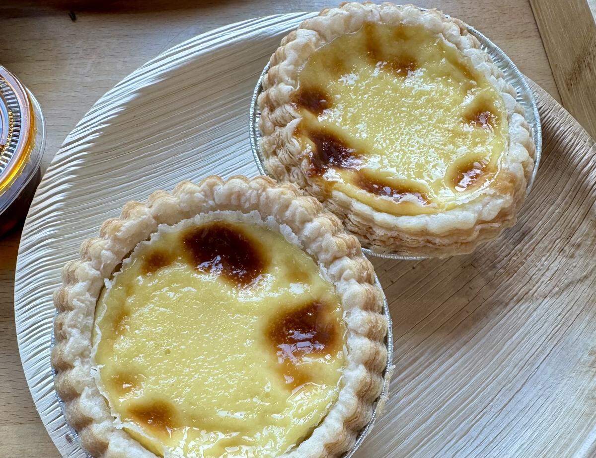 A close-up shot of two egg tarts from Sun’s Kitchen. Each tart sits in a small foil cup and has a circular, flaky crust with a rich yellow custard center. The tops of the custards are browned in spots, giving them a crème brûlée-like appearance. They are presented on a textured, light-brown palm leaf plate.