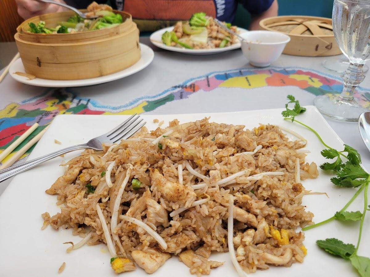 A close-up, eye-level shot of a large, rectangular white plate filled with chicken fried rice. The rice is mixed with pieces of chicken, egg, and fresh bean sprouts, and garnished with cilantro. In the blurred background, other dishes are visible on the table, including a bamboo steamer basket filled with stir-fried vegetables and another plate of food, all resting on a colorful, map-themed paper placemat.