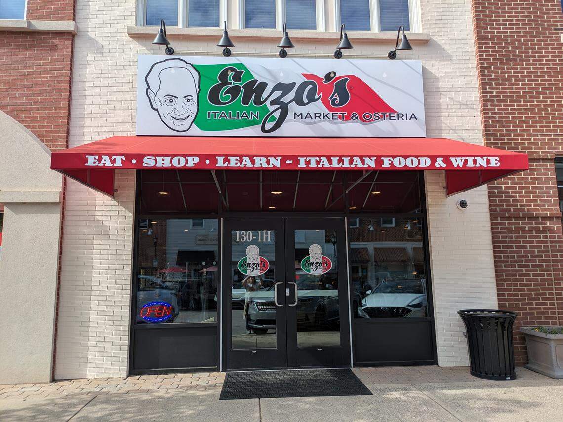 The storefront of “Enzo’s Italian Market & Osteria.” The entrance features black double doors with the company logo on the glass and a red awning above that reads “EAT • SHOP • LEARN ~ ITALIAN FOOD & WINE.” A large sign above the awning displays a caricature of a man’s face next to the brand name in the colors of the Italian flag.