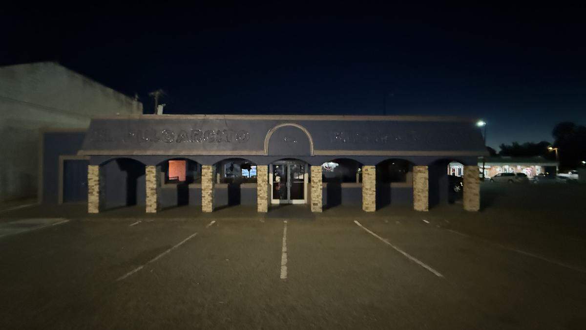 A wide, low-light, nighttime photo of a standalone restaurant with a dark blue facade and a covered front porch supported by several stone-textured columns. The sign above the arched entrance reads “EL PULGARCITO.” The restaurant sits in a large, mostly empty asphalt parking lot with white parking stripes.