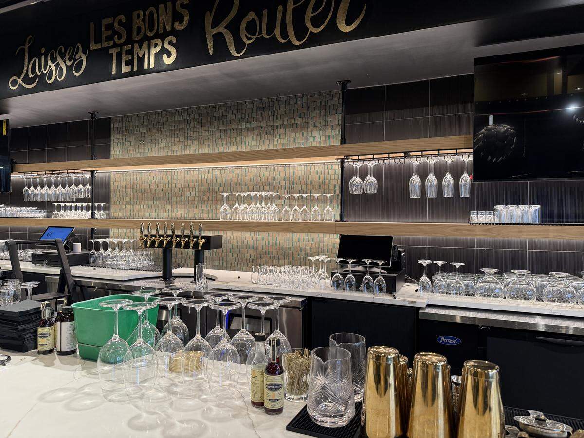 A close-up, high-angle shot of the bar at KCK highlights the sophisticated “behind-the-scenes” details of their beverage service. The foreground features a bright white marble countertop crowded with professional barware, including shimmering gold cocktail shakers, crystal mixing pitchers, and rows of polished wine glasses ready for service. Small bottles of artisanal bitters and syrups are grouped together, suggesting a focus on craft cocktails. A sleek black draft tower with six gold-handled taps sits in the center-left, flanked by a modern point-of-sale (POS) tablet system.