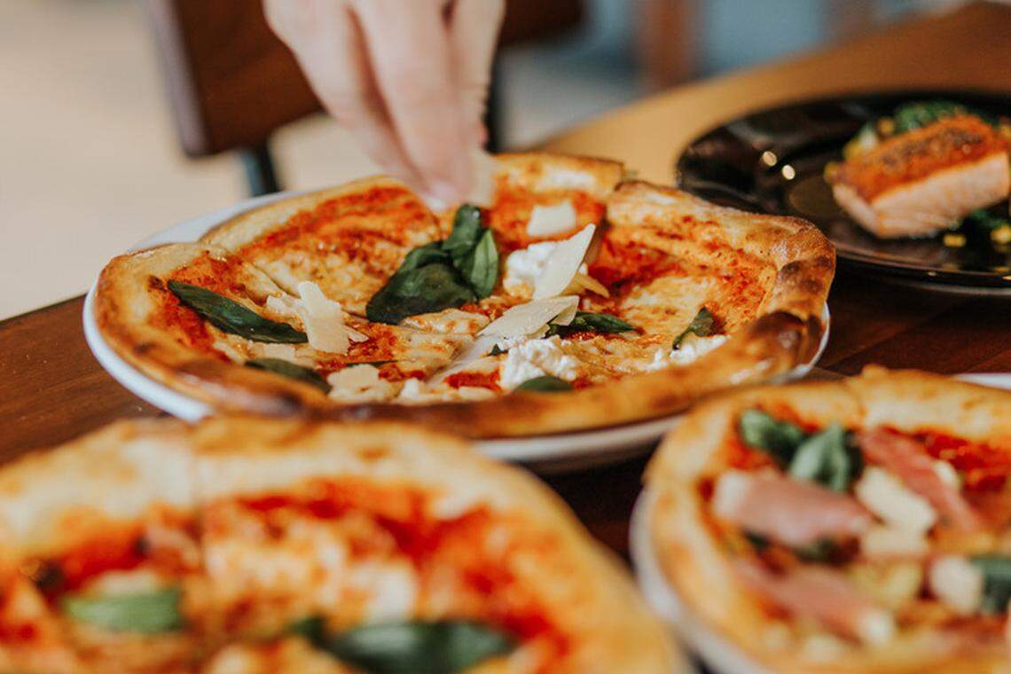 A hand sprinkles cheese over a fresh, round pizza topped with tomato sauce, mozzarella, and basil leaves. The pizza is on a white plate and sits on a wooden table. Other pizzas and a plate with a salmon fillet are visible in the soft-focus foreground and background.