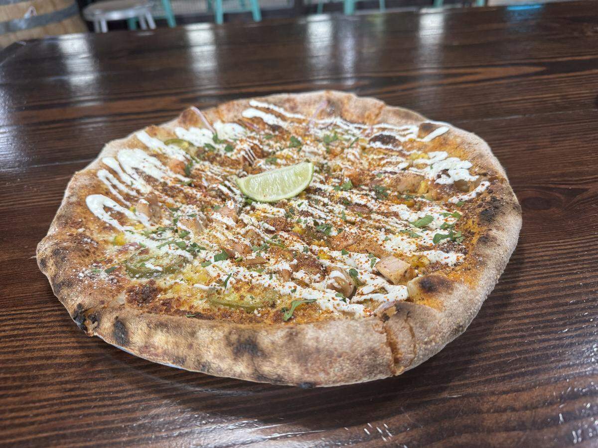 A close-up, overhead shot of a wood-fired pizza with a charred, bubbly crust on a dark wooden table. The pizza is heavily topped with melted cheese, corn, jalapeños, and chicken, drizzled with white crema, and finished with a dusting of red spice, fresh herbs, and a single lime wedge in the center.