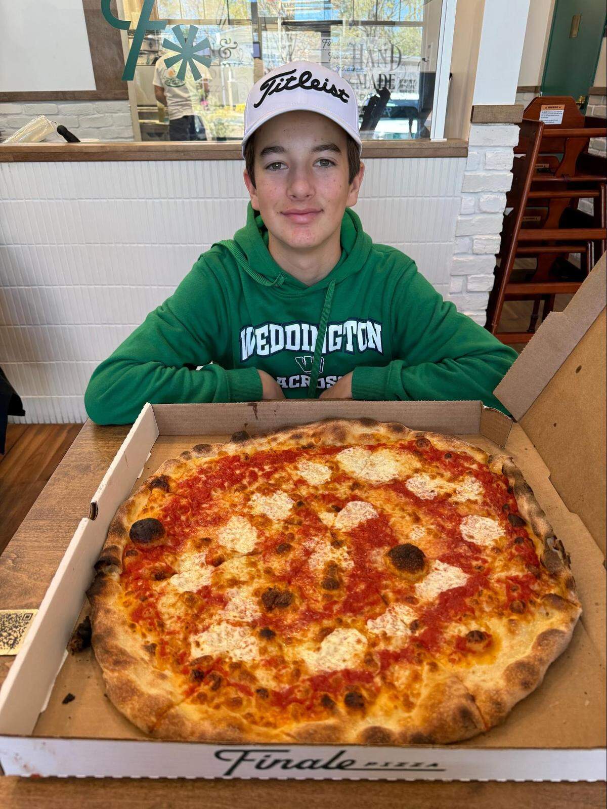 An eye-level, medium shot features a boy wearing a green “WEDDINGTON LACROSSE” hoodie and a white “Titleist” hat, smiling as he sits at a wooden table. In front of him, dominating the foreground, is a large, round cheese pizza with red sauce and dollops of melted mozzarella, resting in an open “Finale” pizza box.