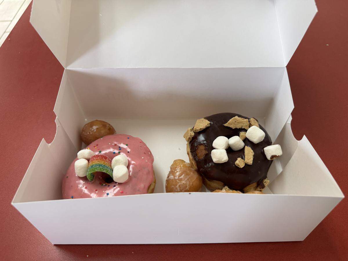 Inside a white cardboard box, there are two specialty doughnuts and several doughnut holes. On the left is a pink-frosted doughnut decorated with small marshmallows and a rainbow sour candy strip. On the right is a chocolate-frosted doughnut topped with marshmallows and crushed graham cracker pieces.