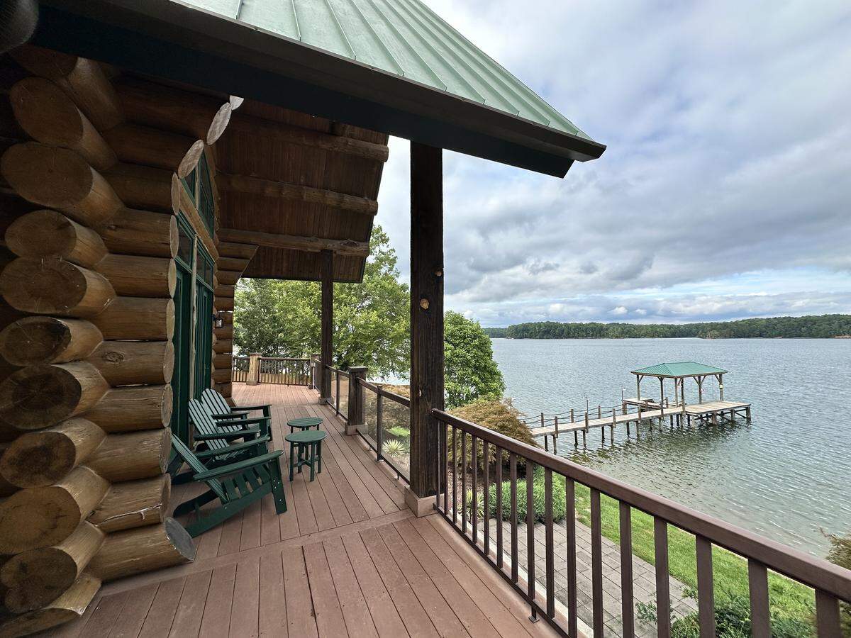 The perspective from the elevated porch of a log cabin, with the large, rounded logs of the house wall on the left. The porch overlooks a wide, calm lake. In the distance, a long dock extends into the water, culminating in a covered structure with a green roof that matches the house. The far shore is covered with trees, and the sky is filled with clouds.