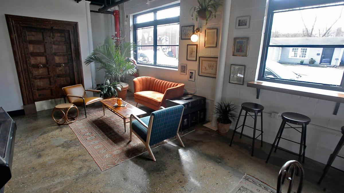 A stylish, industrial-chic cafe or lounge with a cozy seating area. The space has a high ceiling with exposed pipes, white brick walls, and a polished concrete floor. In the center, a small area rug defines a seating arrangement with a low wooden coffee table. The furniture includes a small, pleated orange couch, a light brown leather-look armchair, and a blue plaid armchair. In the background, a large, ornate wooden door with metal details is prominent. The room is decorated with several potted plants, including a large palm, and a gallery wall with various framed pictures. To the right, a long wooden shelf serves as a bar, with two black high-top stools. Large windows let in natural light, with a view of a street and a white house outside.