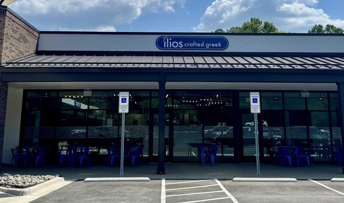 The exterior facade of a contemporary Greek restaurant, “ilios crafted greek,” is shown under a bright, partly cloudy sky. The building features a dark metal awning over a wall of large glass windows and doors, through which blue patio tables and chairs are visible. Two designated accessible parking signs stand prominently in the foreground asphalt parking lot directly in front of the entrance.