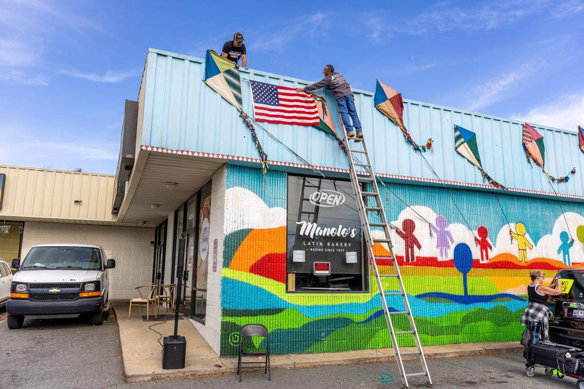 Two men on a ladder affix a large American flag to the blue facade of a Latino-owned bakery in Charlotte. The business owner stands on the roof as the flag is hung. The bakery’s main window is covered in colorful messages and the word “OPEN” is visible on an unlit sign.