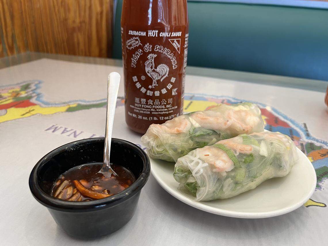 A high-angle, close-up shot of two Vietnamese summer rolls on a small white plate, placed on a map-themed tablecloth. The translucent rolls show shrimp and green herbs inside. Next to the plate is a small black bowl with a dark dipping sauce, and a bottle of Huy Fong Sriracha hot sauce stands in the background.
