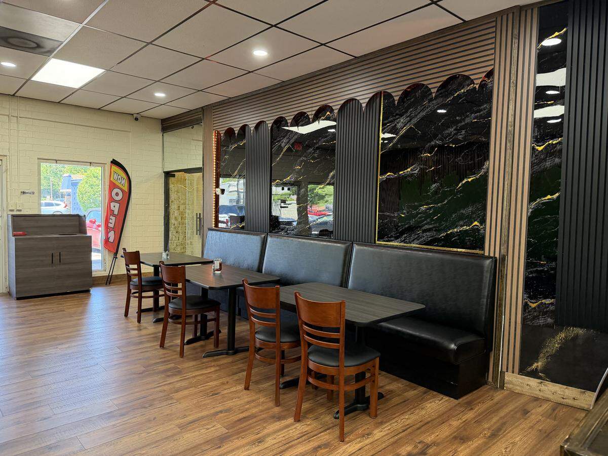 An eye-level shot of a restaurant’s dining area featuring a row of black vinyl booth seating paired with three dark wood laminate tables. Opposite the booths stand four classic wooden dining chairs with black upholstered seats. The accent wall behind the seating features a striking design of black-and-gold marbled panels framed by vertical wooden slats. The space is completed with warm wood-look flooring, a dropped ceiling with recessed lighting, and a red “NOW OPEN” banner visible near a window in the background.