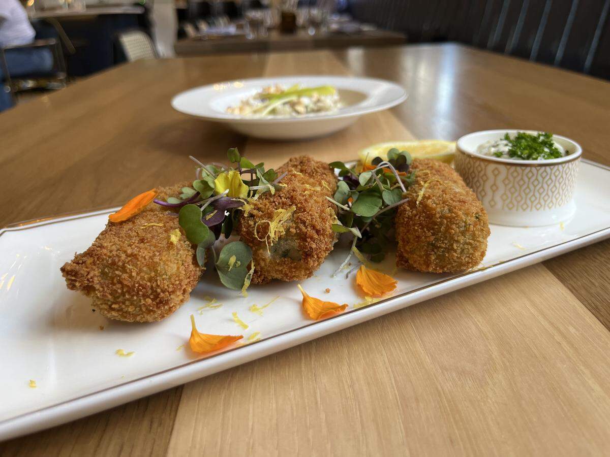 A rectangular white plate with a gold rim holds four golden-brown fried croquettes, garnished with fresh microgreens and orange flower petals. A lemon wedge is tucked among them, and a small white and gold patterned bowl of white dipping sauce, topped with green herbs, sits on the right side of the plate. In the blurred background, another white bowl with a salad or light dish is visible on a wooden table.
