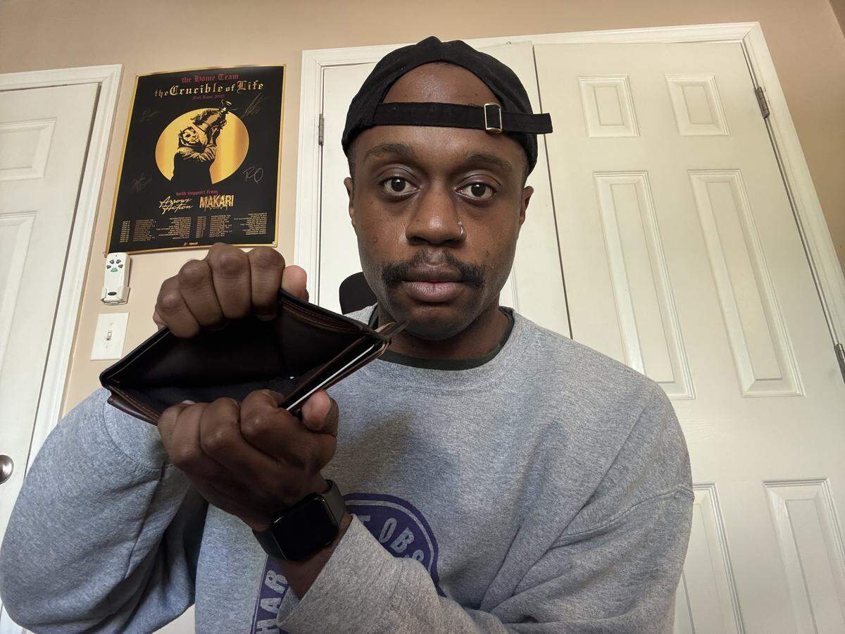 A medium shot of a man with a mustache, wearing a grey sweatshirt and a black baseball cap turned backward, holding open an empty brown leather wallet toward the camera with a deadpan expression.