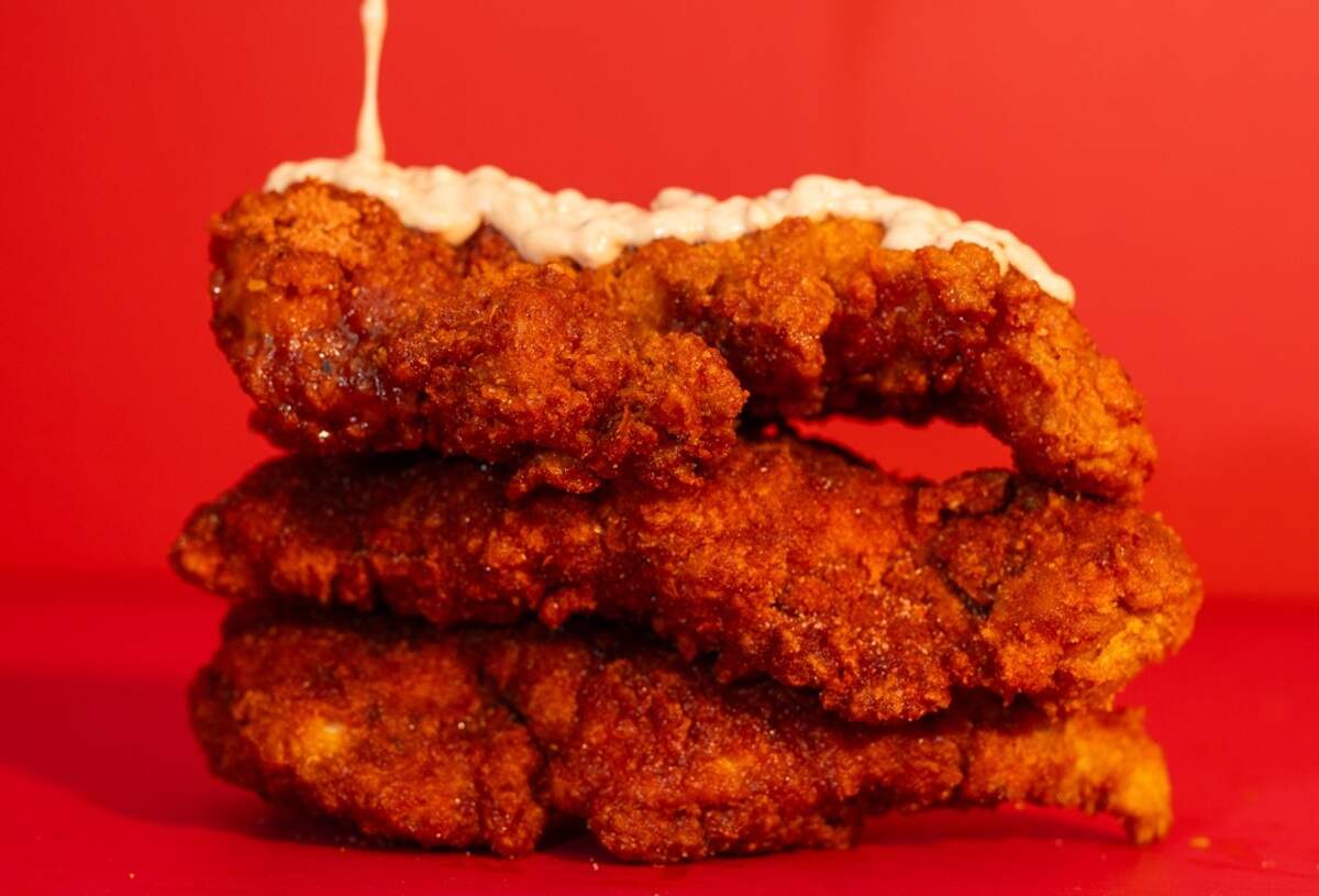 A person is squeezing a creamy white sauce from a bottle onto a stack of three crispy, breaded chicken tenders. The background is a solid red color.