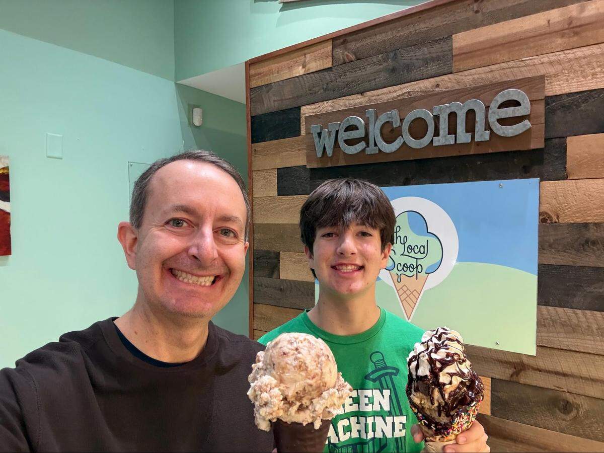 A close-up selfie of two smiling people holding large ice cream treats inside a brightly lit shop. The person on the left is wearing a brown long-sleeved shirt and holds a waffle cone with large scoops of ice cream. The person on the right is wearing a green “GREEN MACHINE” t-shirt and holds a waffle cone rimmed with sprinkles, loaded with ice cream, whipped cream, and chocolate syrup. Behind them is a light green wall featuring a decorative wooden board with the word “welcome” in metal letters, placed above a logo for “The Local Scoop.”