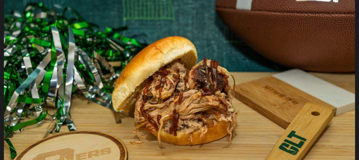 A mouthwatering pulled pork sandwich on a brioche bun with a drizzle of barbecue sauce. In the background, there’s a football and a green and silver cheerleader pom-pom, set against a dark green textured wall. On the wooden table, there are also two wooden coasters, one with a “9ERS” logo and the other a “CLT” logo.