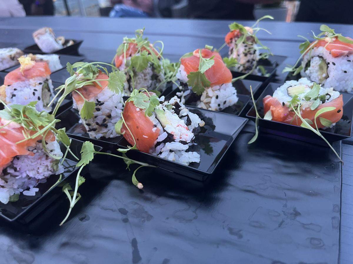 A low-angle shot of a platter of sushi rolls arranged on a dark, wet-looking surface. Multiple pieces of sushi, topped with fresh slices of salmon and green herbs, are presented on small, rectangular black plates. The salmon-topped rolls appear to contain crab and avocado. A few other rolls with different toppings are also visible in the background. The scene is slightly dark, suggesting it might be evening or in a dimly lit setting.
